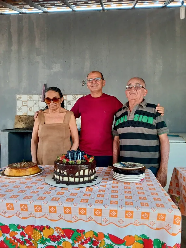 Gilvan de camisa vermelha entre dois familiares, atrás de uma mesa com bolo.