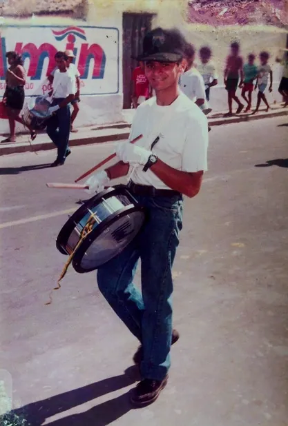 Gilvan jovem, de boné e camisa branca, segurando um tambor em uma rua.
